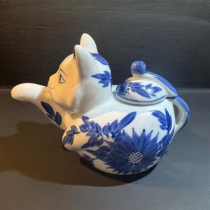 May include: A white and blue ceramic teapot shaped like a cat. The teapot features blue floral designs and a lid. The cat's face is white with blue accents. The teapot has a handle and a spout.