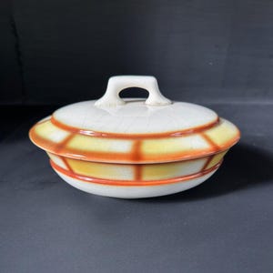 Op de afbeelding: Een vintage keramische schaal met deksel en een witte handgreep. De schaal heeft een wit deksel met een gebarsten glazuur en een geel en oranje geometrisch ontwerp rond de rand. De basis is wit met een oranje streep.