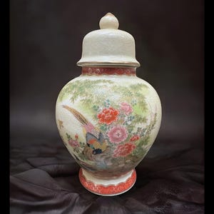 May include: A cream-colored, lidded ceramic vase with a detailed painting of a pheasant and flowers. The vase has a bulbous body, a red and white rim, and a matching lid with a small knob. The artwork features vibrant colors.