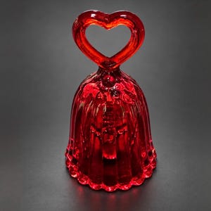 Puede incluir: Una campana de cristal rojo translúcido con un cuerpo acanalado y festoneado, y un asa con forma de corazón abierto. La campana tiene un acabado liso y brillante, con un color rojo claro y vibrante en toda su estructura.