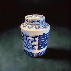 May include: A white and blue ceramic jar with a lid. The jar features blue floral and geometric patterns, along with Chinese characters. The jar is cylindrical with a rounded top and lid, and is likely used for storage.