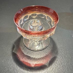 May include: A clear glass candle holder with a red rim. The candle holder has a round base and a detailed design around the bowl. The rim has a decorative pattern. The candle holder is designed to hold a single candle.