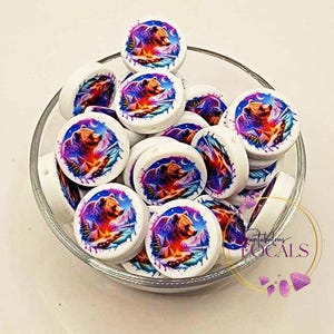 May include: A collection of white, round beads with a colorful bear design inside a glass bowl. The bear illustration features a brown bear, mountains, and a vibrant color palette of purple, orange, and blue. The beads are likely for crafting.
