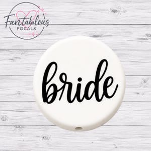 Puede incluir: Una cuenta focal redonda blanca con la palabra "bride" en escritura cursiva negra. La cuenta tiene un pequeño agujero en la parte inferior. El logotipo "Fantabulous Focals" está en la esquina superior izquierda.
