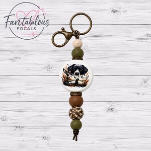 May include: A keychain with a round, white focal bead featuring a black and white dog illustration. The keychain has a bronze-colored clasp and ring, with olive green, brown, and camouflage beads. The text "Fantabulous Focals" is in a circle in the upper left.