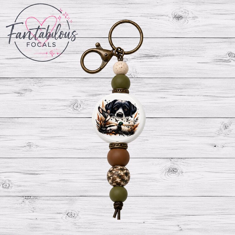 May include: A keychain with a round, white focal bead featuring a black and white dog illustration. The keychain has a bronze-colored clasp and ring, with olive green, brown, and camouflage beads. The text "Fantabulous Focals" is in a circle in the upper left.