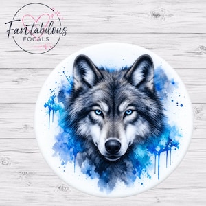 May include: A circular decorative item featuring a detailed illustration of a wolf head with piercing blue eyes. The wolf is surrounded by vibrant blue and dark grey watercolor-style paint splatters and drips on a clean white background.