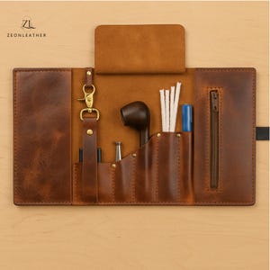 Puede incluir: Estuche de cuero marrón abierto para pipa de tabaco. El estuche contiene una pipa, herramientas de limpieza y un bolígrafo azul. Se ven una cremallera y un cierre. La marca "ZEONLEATHER" está impresa en la parte superior izquierda.