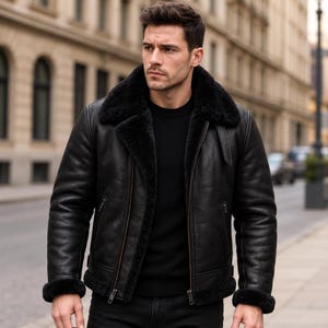 Op de afbeelding: Een zwart leren jack met een dikke zwarte shearling kraag, binnenvoering en manchetten. Het jack is voorzien van een ritssluiting aan de voorzijde, twee zijzakken met rits en een gespdetail op de kraag, gedragen over een zwarte trui.