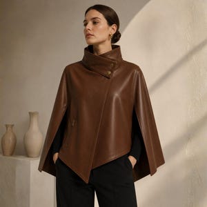 May include: A brown leather cape with a high collar and asymmetrical front closure. The cape features a zipper pocket and two gold-colored buttons. The model is wearing black pants and a black top.