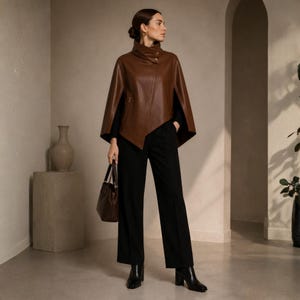 May include: A brown leather cape with a high collar and button closure. The cape is paired with black pants and black heeled boots. A brown handbag is held in the hand. The background is a neutral color.
