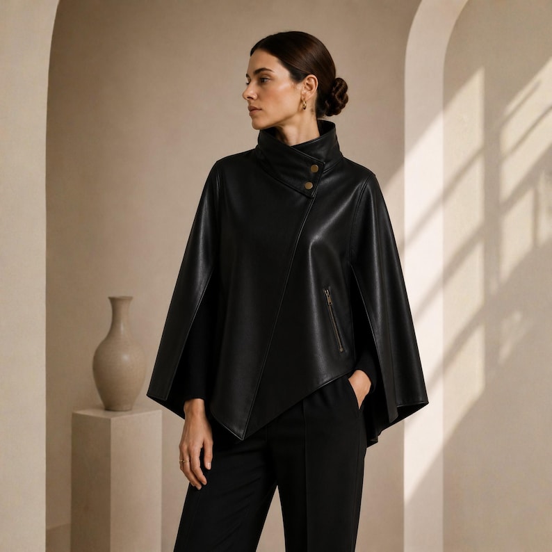 May include: A black leather cape with a high collar and asymmetrical front. The cape features two gold-colored buttons and a zippered pocket. The model is wearing black pants and a black top.