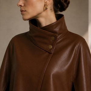 May include: A brown leather cape with a high, rounded collar and two gold-colored buttons. The cape has a smooth texture and a slight sheen, suggesting a high-quality material. The design is modern and elegant, suitable for outerwear.