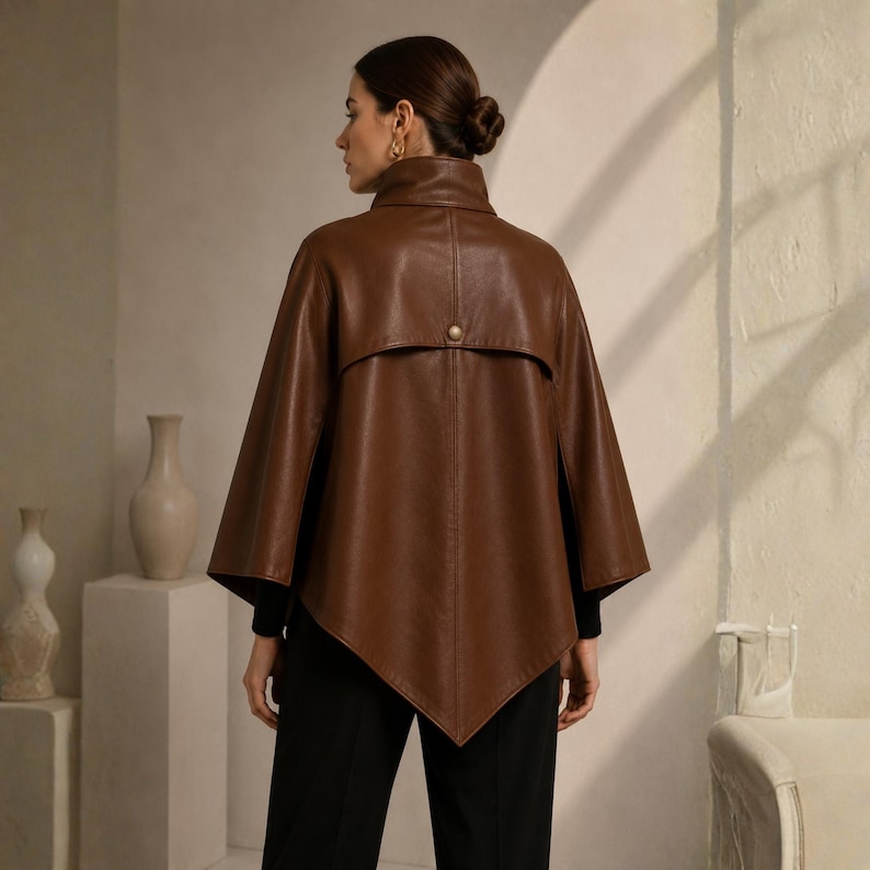 May include: A brown leather cape with a high collar and a single button closure. The cape has a unique, angular hemline and is worn over a black top and pants. The overall style is sophisticated and chic.