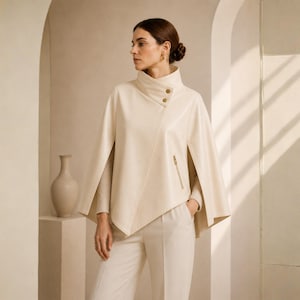 May include: Cream-colored cape jacket with a high collar and asymmetrical design. The jacket features gold-toned buttons and a zipper pocket. The model is wearing matching cream-colored pants. A neutral-toned vase is in the background.