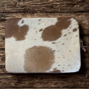 May include: Rectangular leather laptop sleeve with a cowhide pattern. The sleeve has a zip closure and a brown and white spotted design. The brand name "ZEONLEATHER" is visible in the top left corner.