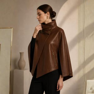 May include: A brown leather cape with a high collar and asymmetrical front. The cape features a zipper detail and button closures. The model is wearing a black top and black pants. The cape is a stylish outerwear option.