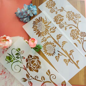 May include: White floral stencils featuring sunflower, rose, and other botanical designs. The stencils are made of a flexible material and are ideal for various craft projects. The image also includes artificial flowers in pink and blue.