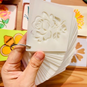 Könnte beinhalten: Ein Satz weißer Kunststoff-Schablonen mit verschiedenen Designs, darunter eine Rose, ein Schmetterling, eine Biene, ein Herz und eine Blume. Die Schablonen werden durch einen silbernen Ring zusammengehalten.