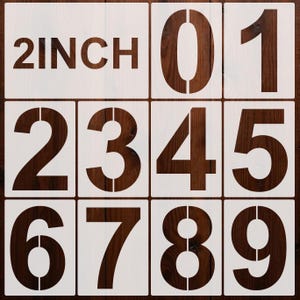Op de afbeelding: Negen witte sjablonen met de cijfers 0 tot en met 9, en een sjabloon met de tekst "2 INCH". De sjablonen zijn geplaatst op een donkerbruine houten achtergrond. De cijfers zijn in een vetgedrukte, schreefloze lettertype.