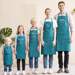 May include: A group of people wearing teal aprons with white text. The aprons have a pocket and are personalized with names and titles like "Chef" and "Chef in Training." The aprons are worn by people of different ages, suggesting a family or group activity.
