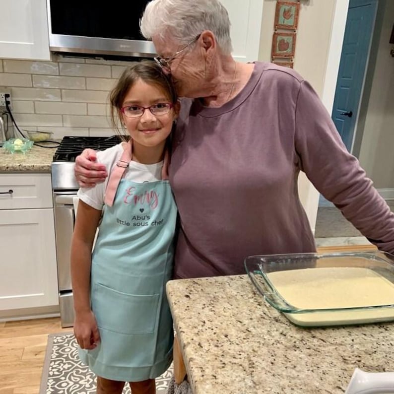 Pu&ograve; includere: Una bambina indossa un grembiule azzurro con la scritta "Emily" e "Abu's little sous chef" in una cucina. Una donna anziana la abbraccia. Una pirofila di vetro con una sostanza gialla &egrave; sul bancone. Scena di cucina.