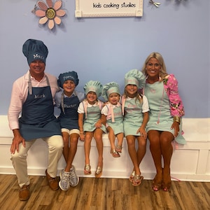 Pu&ograve; includere: Un gruppo di sei persone che indossano grembiuli e cappelli da chef abbinati, in posa davanti a un cartello con la scritta "flour power kids cooking studios". I grembiuli e i cappelli sono blu navy e azzurri. Il cartello ha un logo dello chef.