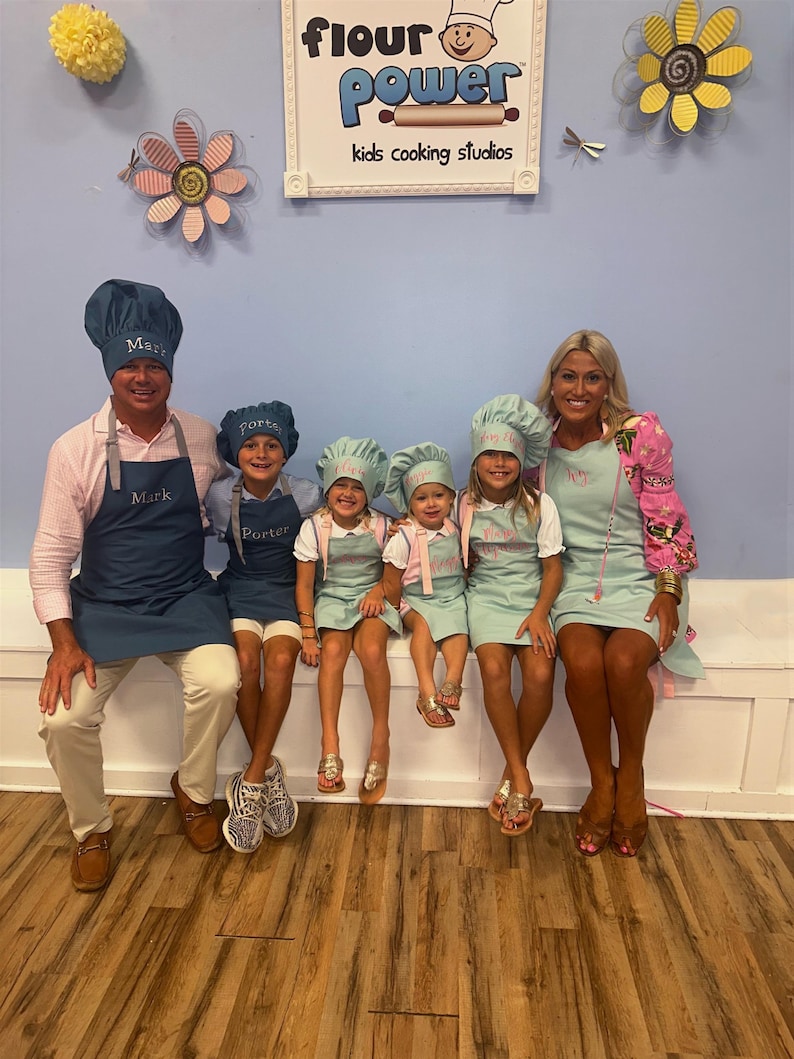 Pu&ograve; includere: Un gruppo di sei persone che indossano grembiuli e cappelli da chef abbinati, in posa davanti a un cartello con la scritta "flour power kids cooking studios". I grembiuli e i cappelli sono blu navy e azzurri. Il cartello ha un logo dello chef.