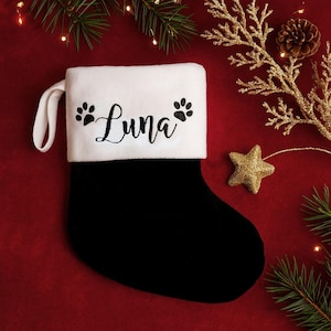 May include: A black and white Christmas stocking with the name "Luna" embroidered in black script. The stocking has paw prints on either side of the name. A white loop is attached to the top. The stocking is on a red surface with Christmas decorations.