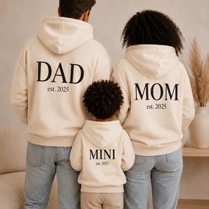Könnte beinhalten: Drei beige Kapuzenpullover mit schwarzem Text auf der Rückseite. Der größte Hoodie zeigt DAD est. 2025, der zweite MOM est. 2025 und der kleinste MINI est. 2025. Jedes Kleidungsstück verfügt über eine Kapuze und lange Ärmel.