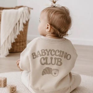 Könnte beinhalten: Ein beigefarbenes Langarm-Sweatshirt mit dem Schriftzug BABYCCINO CLUB in geschwungener brauner Schrift auf der Rückseite. Unter dem Text befindet sich eine grafische Darstellung eines Croissants und einer kleinen Tasse mit Herzmotiv.