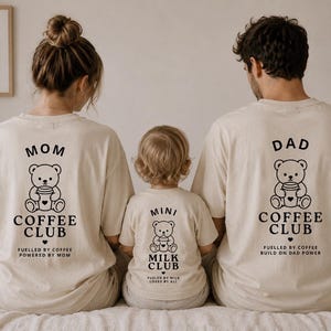 Könnte beinhalten: Drei beige T-Shirts mit schwarzen Grafikdrucken. Die beiden größeren Shirts zeigen einen Bären mit Kaffeetasse und den Text MOM COFFEE CLUB sowie DAD COFFEE CLUB. Das kleinere Shirt zeigt einen Bären mit dem Text MINI MILK CLUB.