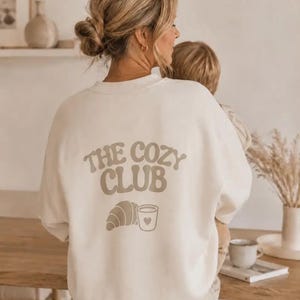 Könnte beinhalten: Ein cremefarbenes Sweatshirt mit Rundhalsausschnitt und dem Schriftzug THE COZY CLUB in einer großen, geschwungenen, beigefarbenen Schrift auf der Rückseite. Unter dem Text befindet sich eine Grafik eines Croissants und einer Kaffeetasse.