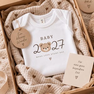 Könnte beinhalten: Ein weißer Babybody mit dem Text BABY 2027 und einer Bärenillustration sowie dem Schriftzug MORE LOVE EVERY DAY. Dazu gehören ein rundes Etikett mit der Aufschrift Ein kleines Wunder, ein beiges Musselintuch mit braunen Herzen und eine Karte Für eine ganz besondere Zeit.