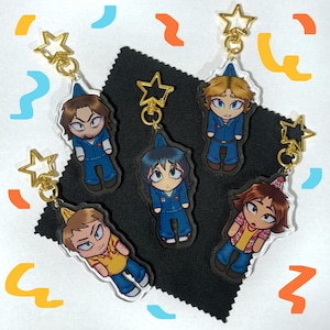 以下が含まれることがあります： 金色の星のチャームが付いた、カラフルな漫画のキャラクターのキーホルダーが5つ。それぞれのキーホルダーには、ユニークな表情と服装をした異なるキャラクターが描かれています。キャラクターは青と黄色の服を着ています。