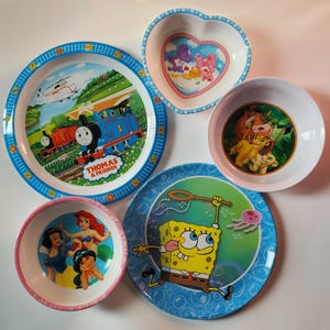 May include: A collection of children's plates and bowls featuring cartoon characters. The plates have blue rims and depict Thomas the Tank Engine, Spongebob Squarepants, and other characters. The bowls feature Care Bears, Ariel, and Simba.