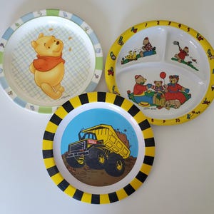 May include: Three colorful plastic children's plates. One plate features Winnie the Pooh, another has a dump truck, and the third has a divided section with bears and numbers 1-10.