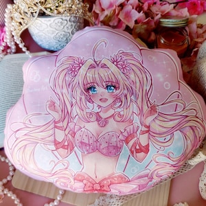 Puede incluir: Una almohada rosa y blanca con un diseño de concha. La almohada presenta una ilustración de dibujos animados de una niña con el pelo largo y rubio que lleva un top de bikini rosa y blanco de concha. La niña está rodeada de purpurina rosa y blanca.