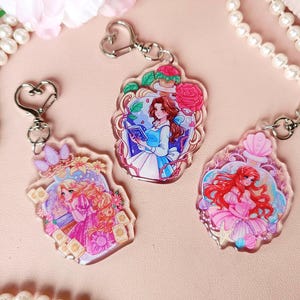 Pode incluir: Três chaveiros coloridos com princesas da Disney. Cada chaveiro tem uma princesa diferente: Bela, Ariel e Rapunzel. Os chaveiros são feitos de acrílico transparente e têm uma corrente de metal prateada.