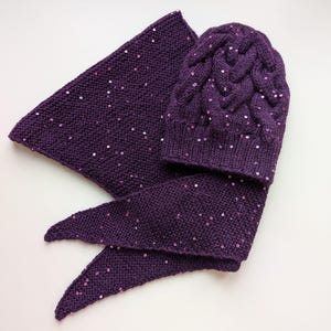 May include: A purple knitted hat and scarf set with sparkly sequins. The hat has a cable knit pattern and the scarf is a triangular shape.