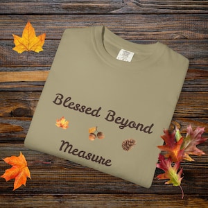 May include: A khaki-colored t-shirt with the words "Blessed Beyond Measure" in brown script. The shirt features illustrations of a leaf, acorns, and a pinecone. Autumn-themed apparel.
