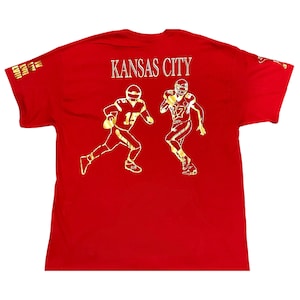 Può includere: T-shirt rossa con grafica dorata di due giocatori di football americano in azione. La scritta "KANSAS CITY" è sopra i giocatori. La maglietta ha anche del testo sulle maniche. Per i fan della squadra di football di Kansas City.