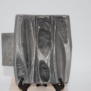 Puede incluir: Una placa rectangular de metal gris con una superficie texturizada e irregular que presenta profundas ranuras verticales y hendiduras oscuras en forma de cráter. El objeto tiene una sección rectangular plana que sobresale en el lado izquierdo.