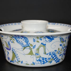 May include: A white ceramic casserole dish with a lid, featuring a blue and green line-art illustration of two figures sitting among foliage and birds. The bowl has a wide, flared rim with integrated handles and a central knob on the lid.