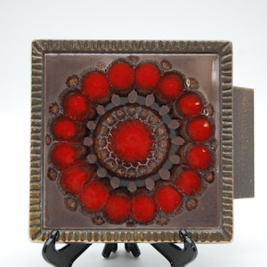 May include: Square ceramic tile with a brown and red floral design. The tile features a central red circle surrounded by red, circular shapes. The tile is framed with a textured, brown border. The tile is displayed on a black stand.