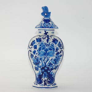 Könnte beinhalten: Ein blau-weißes Keramik-Ingwerglas mit Blumenmuster und einem Deckel, der von einer kleinen, geformten Tierfigur gekrönt wird. Das Gefäß hat einen bauchigen Körper, der sich zu Basis und Hals hin verjüngt und mit blauen Blumenmotiven verziert ist.