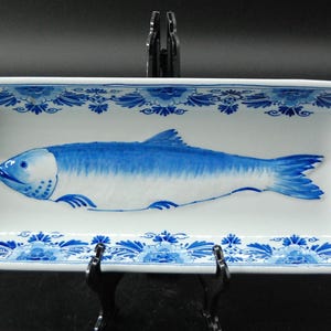 Könnte beinhalten: Eine rechteckige Servierschale aus weißer Keramik mit einem handgemalten blauen Fisch in der Mitte. Der innere Rand ist mit einem sich wiederholenden blauen Blumenmuster verziert. Die Schale steht auf einem schwarzen Kunststoffständer.