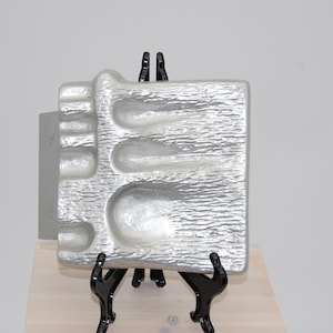 May include: A silver-coloured, textured decorative tray with rounded indentations, displayed on a black easel. The tray has a rectangular shape with a unique, organic design. The easel is made of black metal.