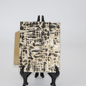 May include: A rectangular cuff bracelet featuring a textured surface with a high-contrast pattern of raised metallic silver elements and recessed black grooves. The bracelet has a smooth, polished gold-tone interior band visible on the side.