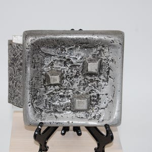 May include: A square, metallic gray tray with a heavily textured, pitted surface. The tray features three raised, smooth, square-shaped accents positioned asymmetrically on its face. The object is displayed on a black, three-legged stand.